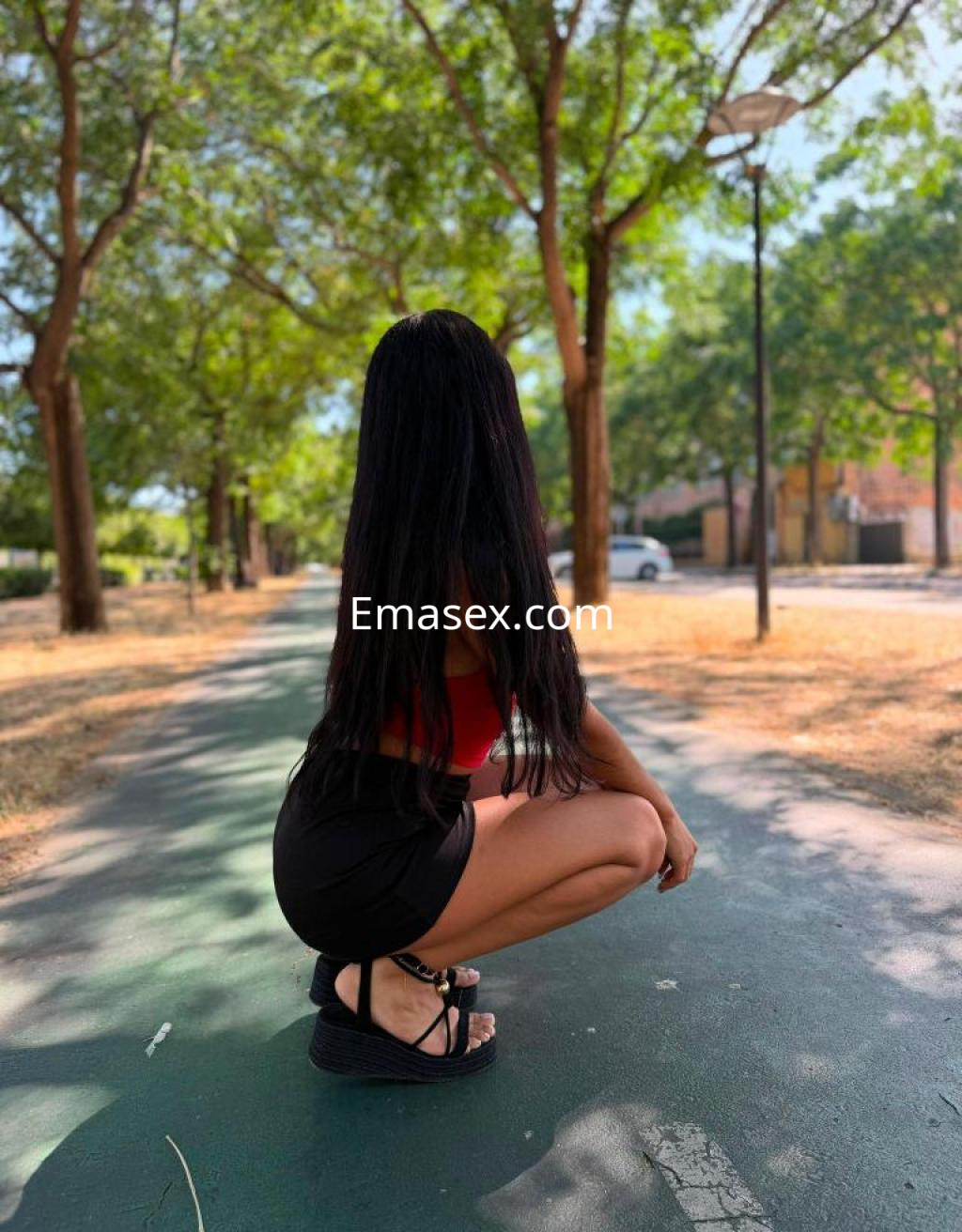 Puta en Sevilla, agachada sobre un camino de parque, con cabello largo y oscuro, usando top rojo y short negro.