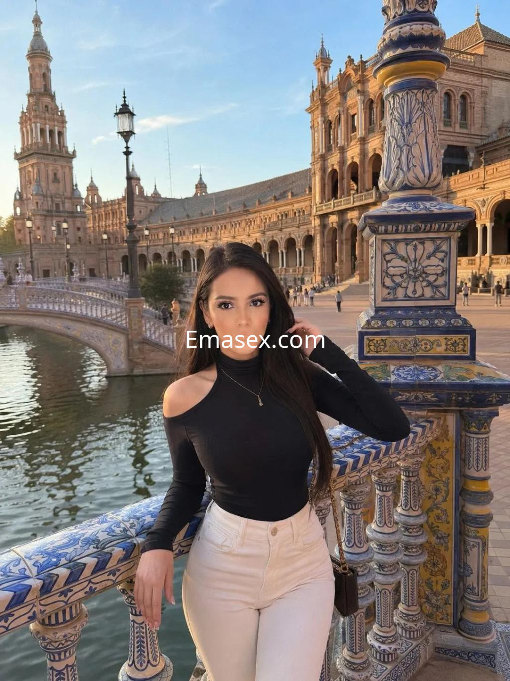 Puta posando con un top negro y pantalones claros en la Plaza de España de Sevilla, con un paisaje arquitectónico.