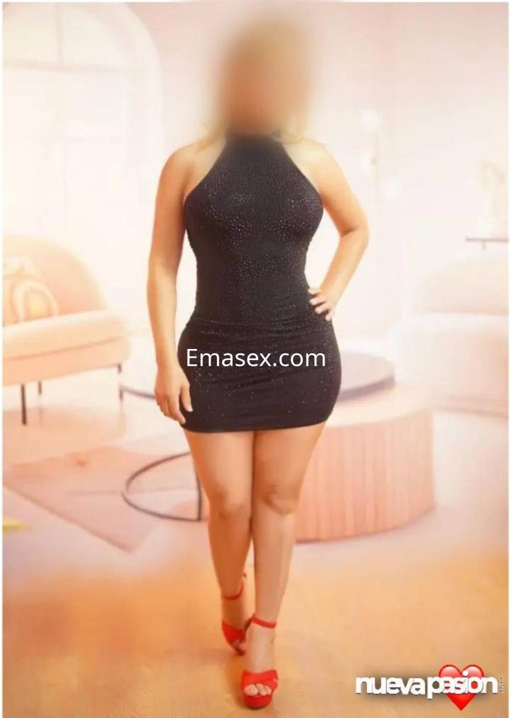 Puta en Sevilla con un vestido negro ajustado y sandalias rojas, posando en un ambiente acogedor.