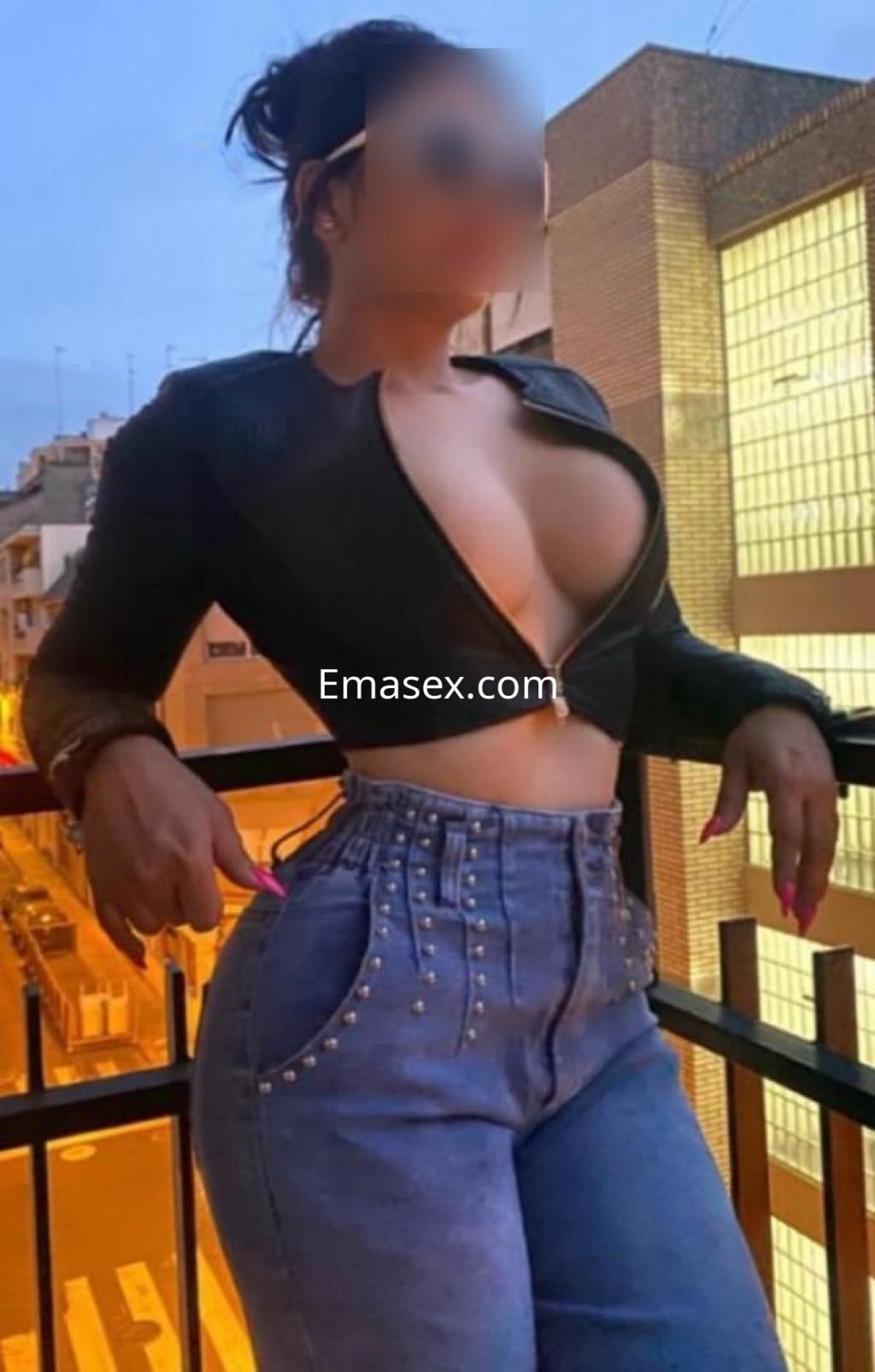 Escort en Tarragona posando en un balcón con blusa negra ajustada y jeans de tiro alto, luciendo segura.