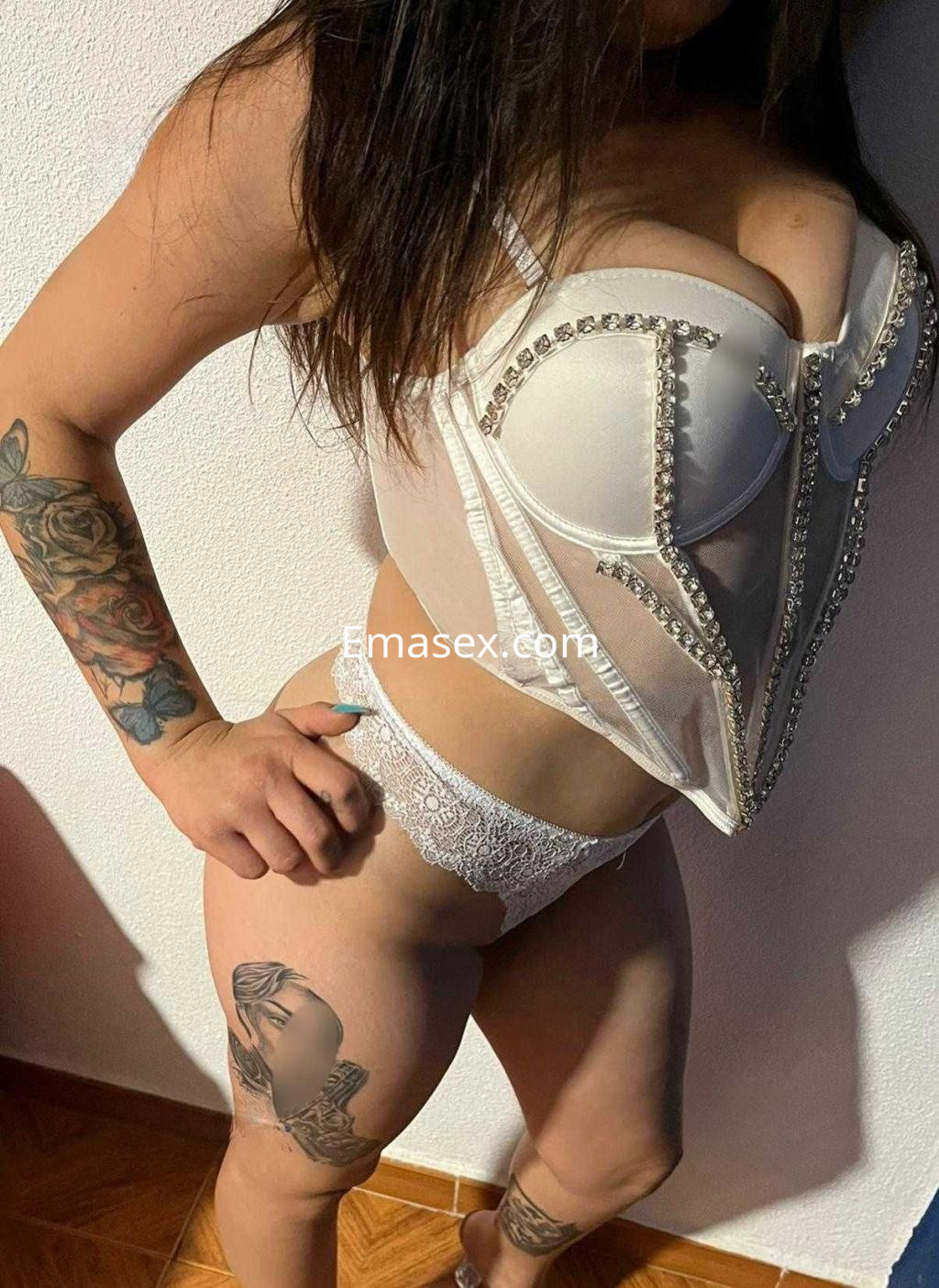 Escort en Valladolid posando con un corsé blanco decorado y lencería, mostrando tatuajes en su muslo.