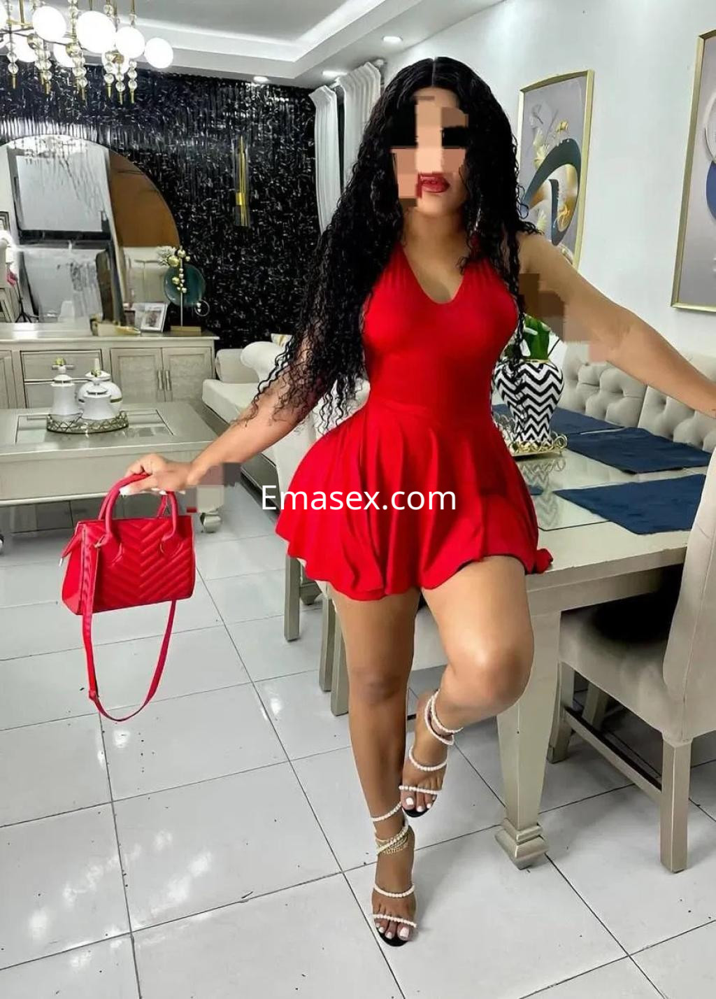 Puta en Valladolid viste un vestido corto rojo y sandalias de tacón, posando junto a una mesa moderna.