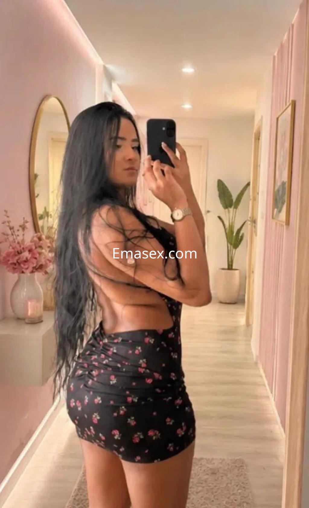 Escort en Zaragoza con cabello largo y lacio, viste un vestido negro sin mangas con estampado floral y se toma una selfie.