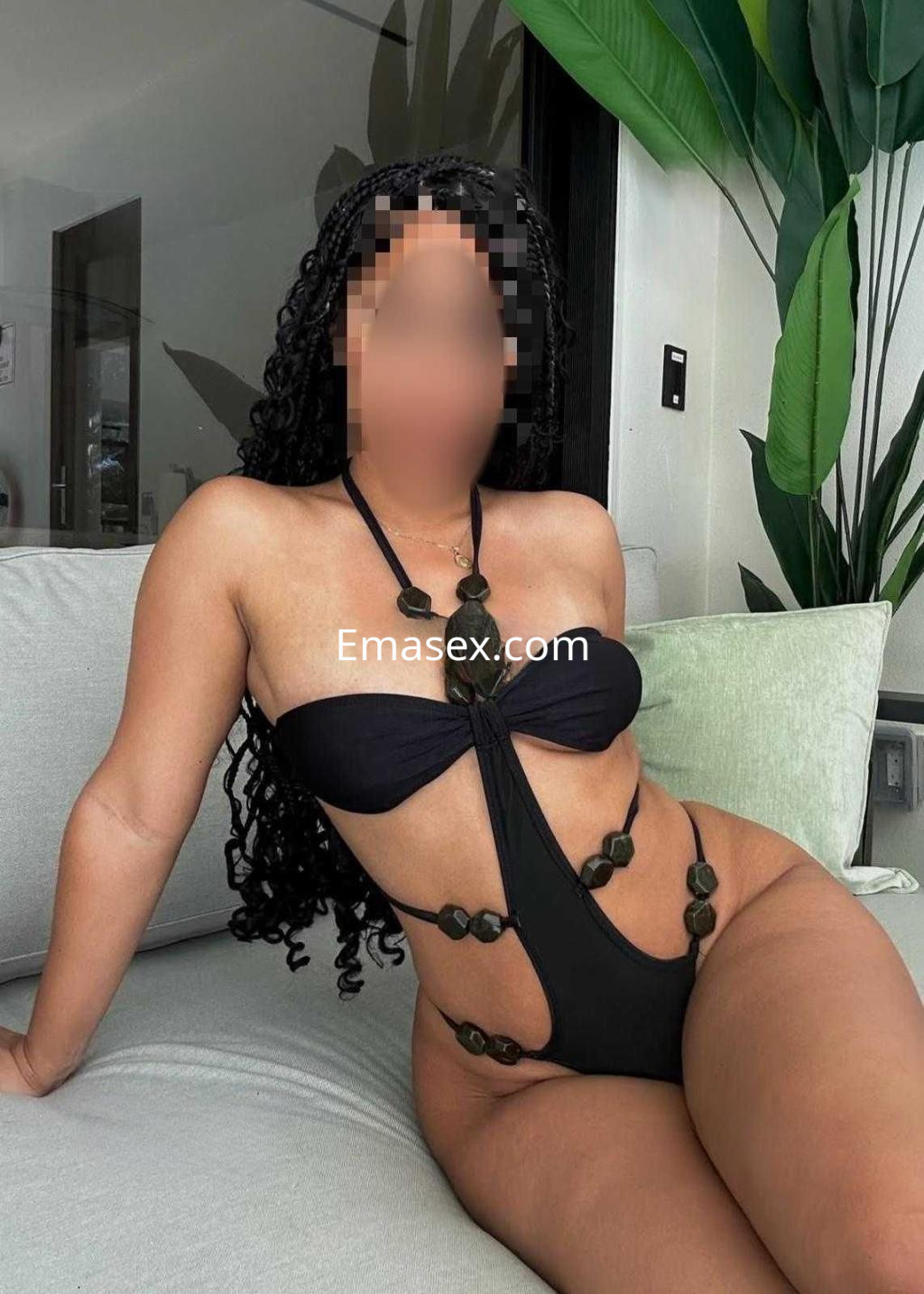 Puta posando en Zaragoza con un bikini negro, sentada en un sofá y rodeada de plantas decorativas.