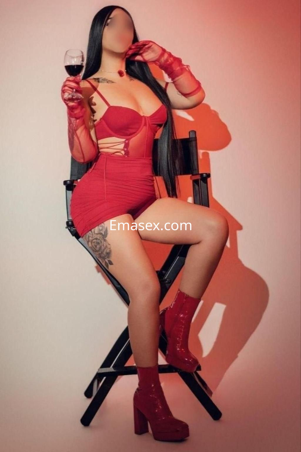 Puta en Almería luciendo un vestido rojo ajustado y botas altas, posando con una copa de vino en mano.