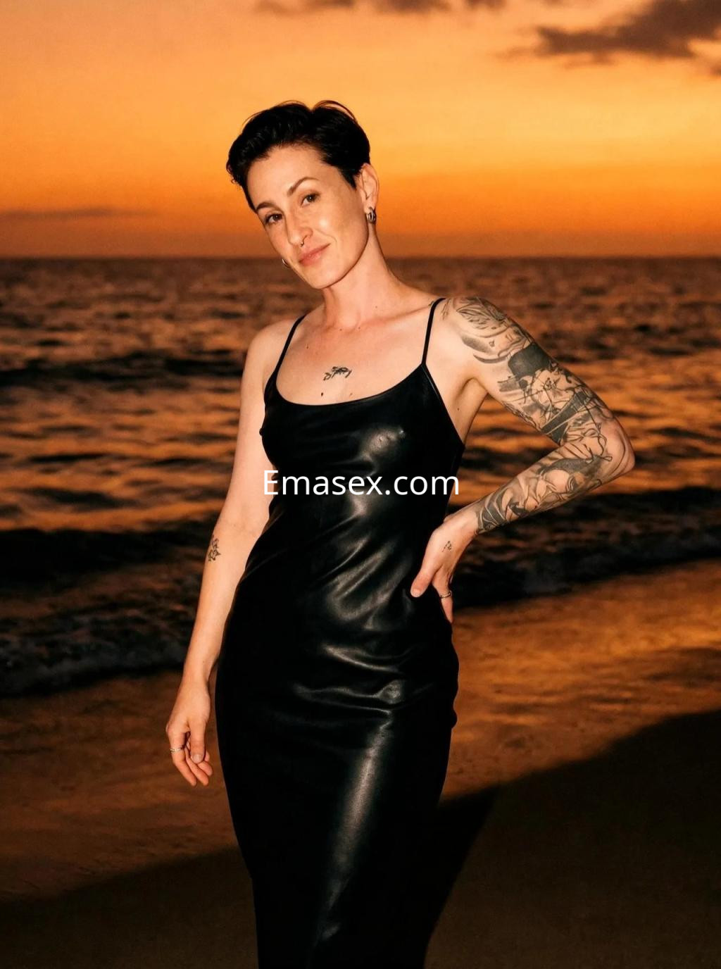Escort en Málaga, posando en la playa al atardecer, con un vestido negro ajustado y tatuajes visibles.