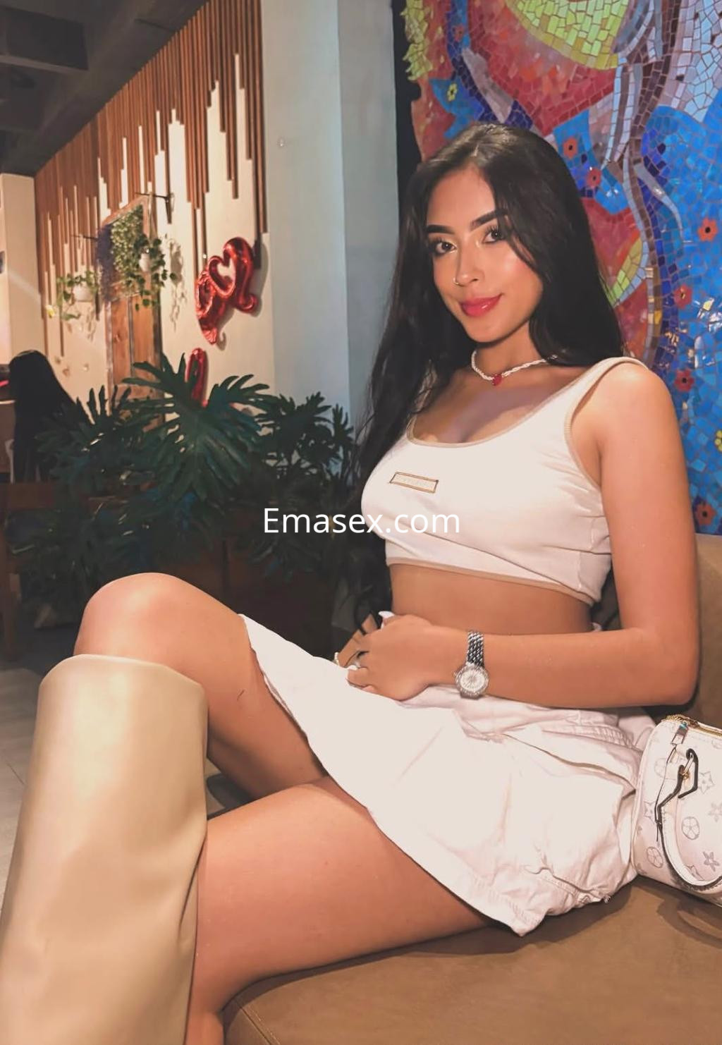Escort en Pamplona con cabello largo y lacio, luciendo un top blanco y una falda corta, sentada en un ambiente moderno.