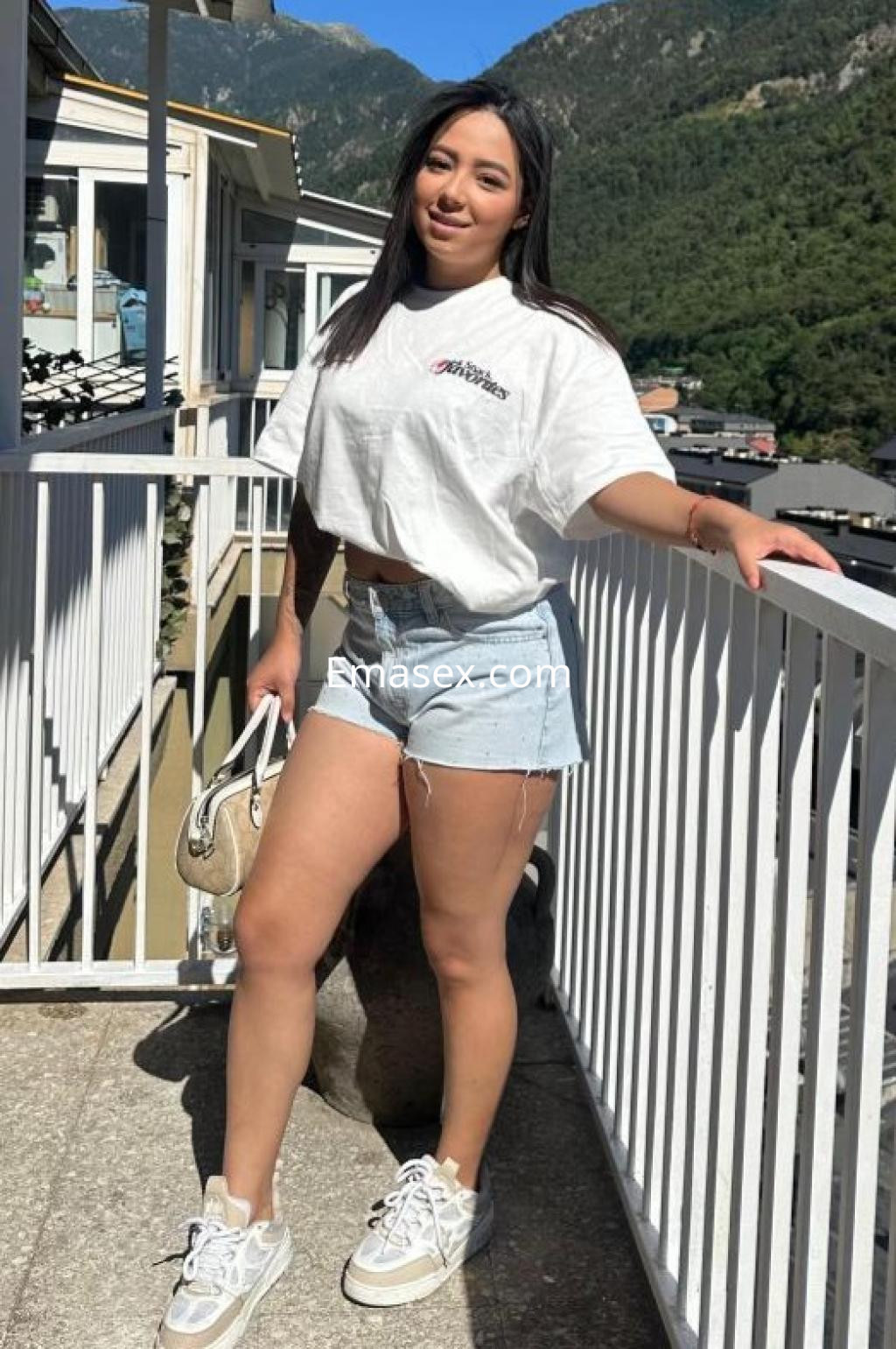 Escort en Lleida con camiseta blanca y shorts de mezclilla, posando al aire libre con un paisaje montañoso de fondo.