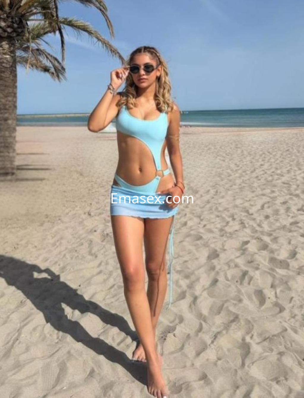 Puta en Lleida, luciendo un vestido de baño azul claro y una falda corta en la playa, con gafas de sol.