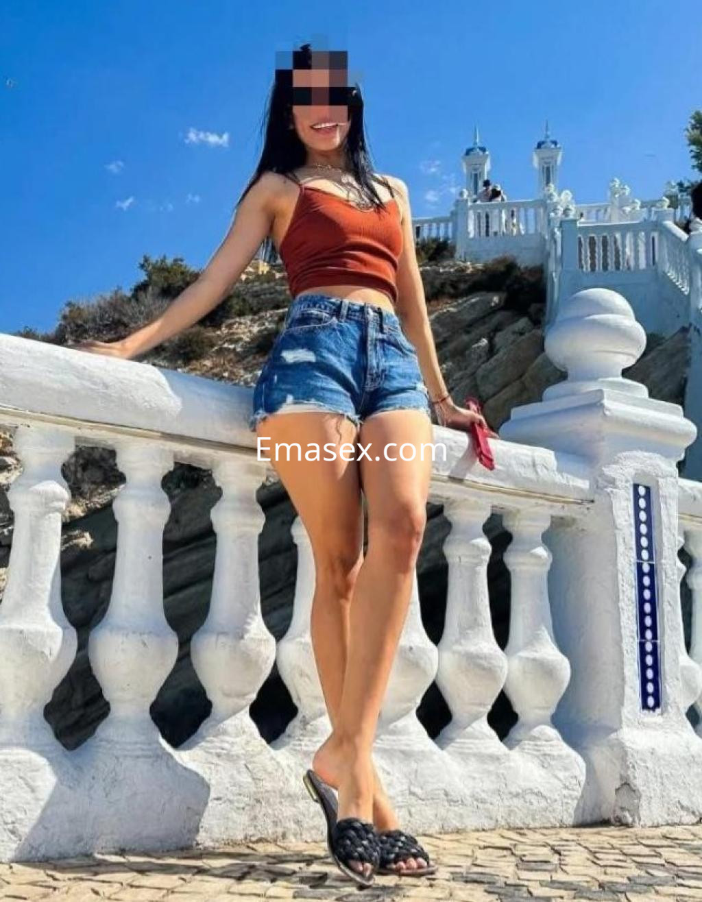Escort en Girona, de pie junto a una barandilla, lleva un top marrón y pantalones cortos, con una sonrisa y gafas de sol.