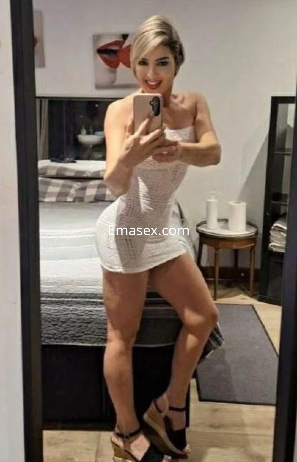 Puta posando frente al espejo en Girona, vistiendo un vestido blanco ajustado y sandalias de tacón.