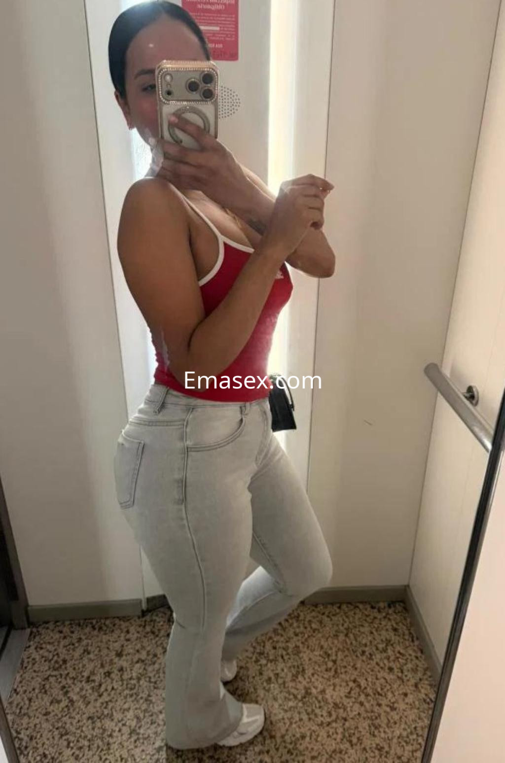 Escort en Castellón posando frente al espejo con una blusa roja y jeans ajustados, mostrando una sonrisa.