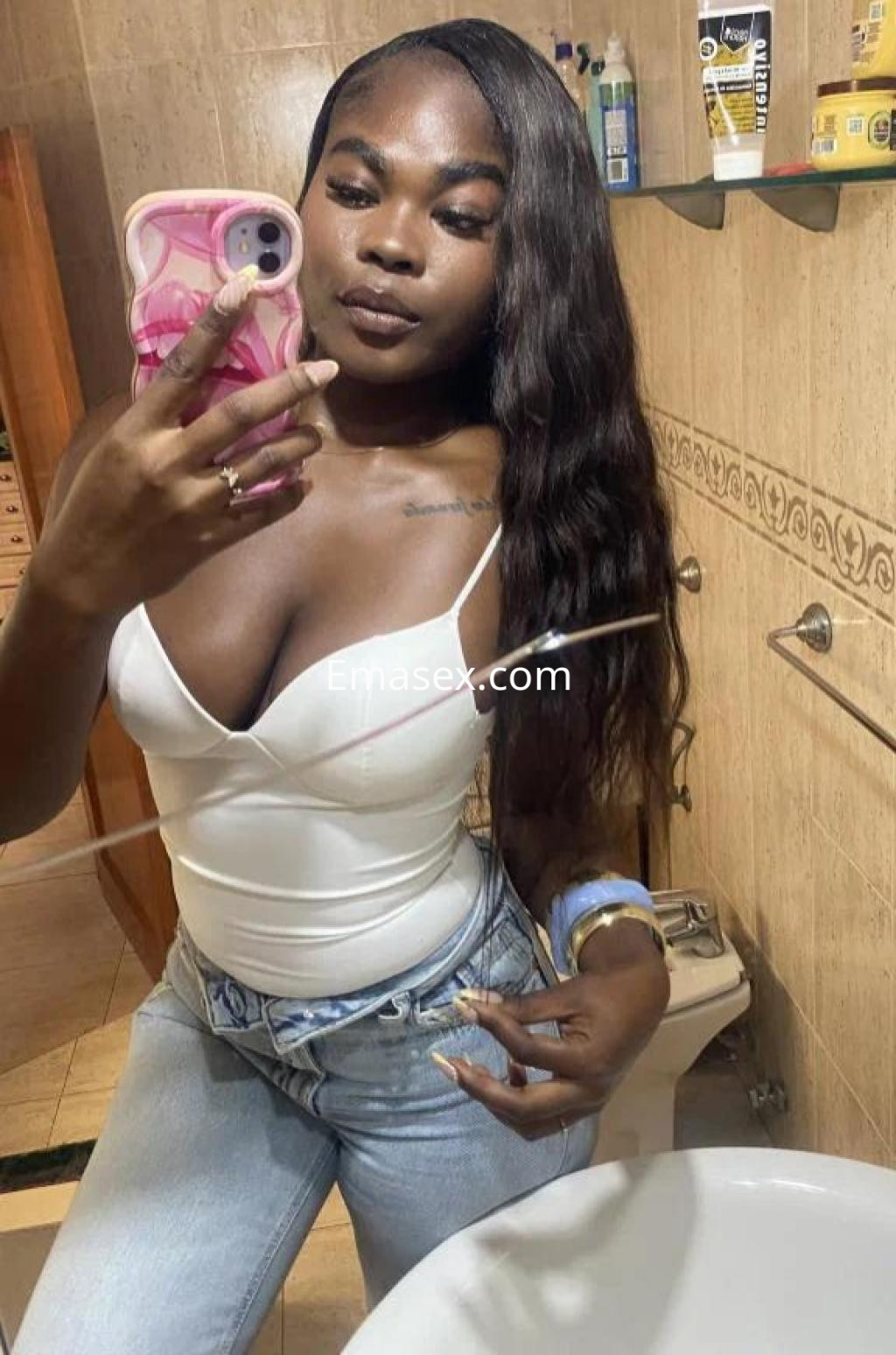 Puta en Las Palmas posando en un baño, viste una blusa blanca ajustada y jeans, con cabello largo y oscuro.