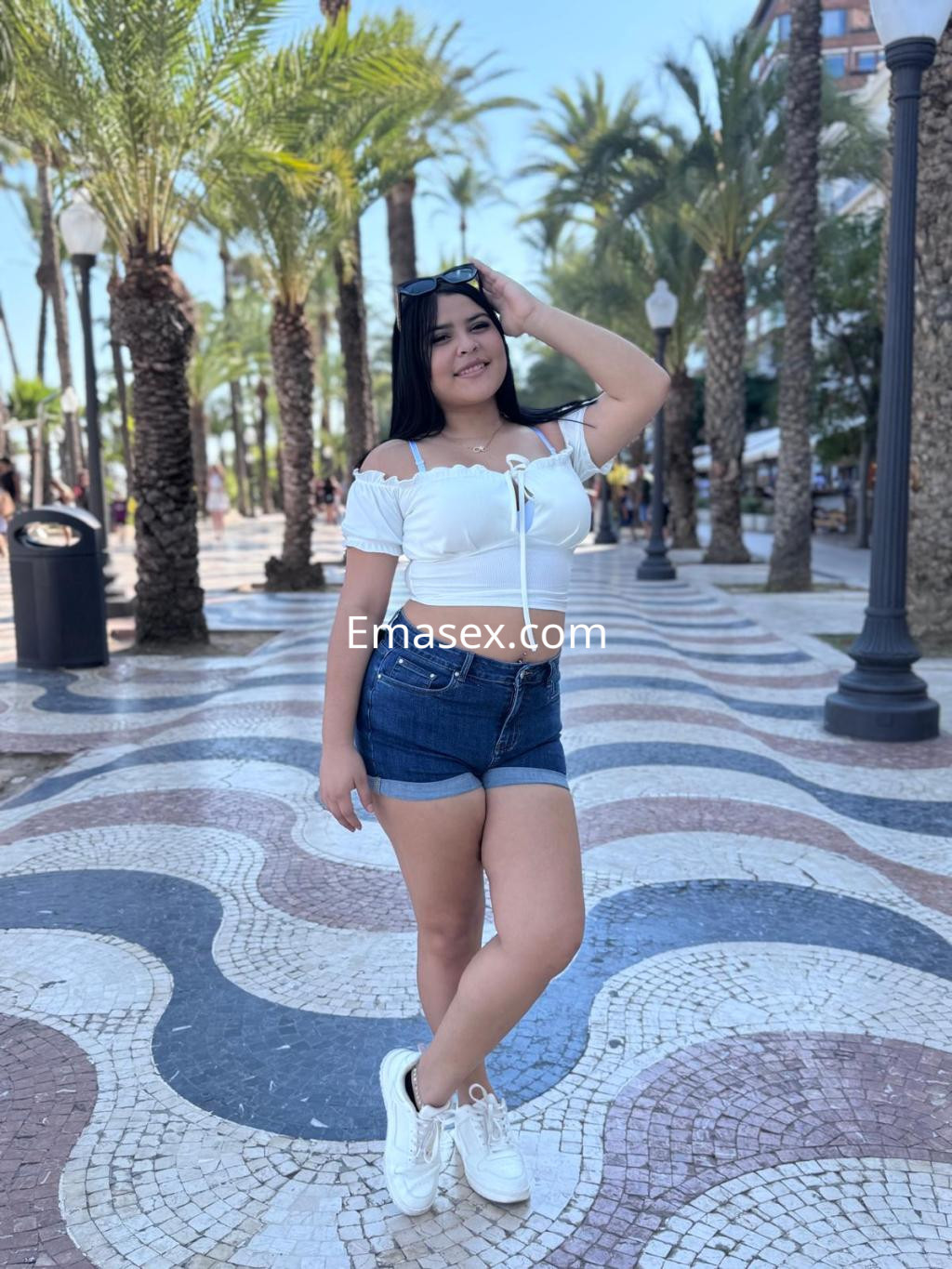 Escort en Alicante con blusa blanca y pantalones cortos, posando entre palmeras en un paseo.