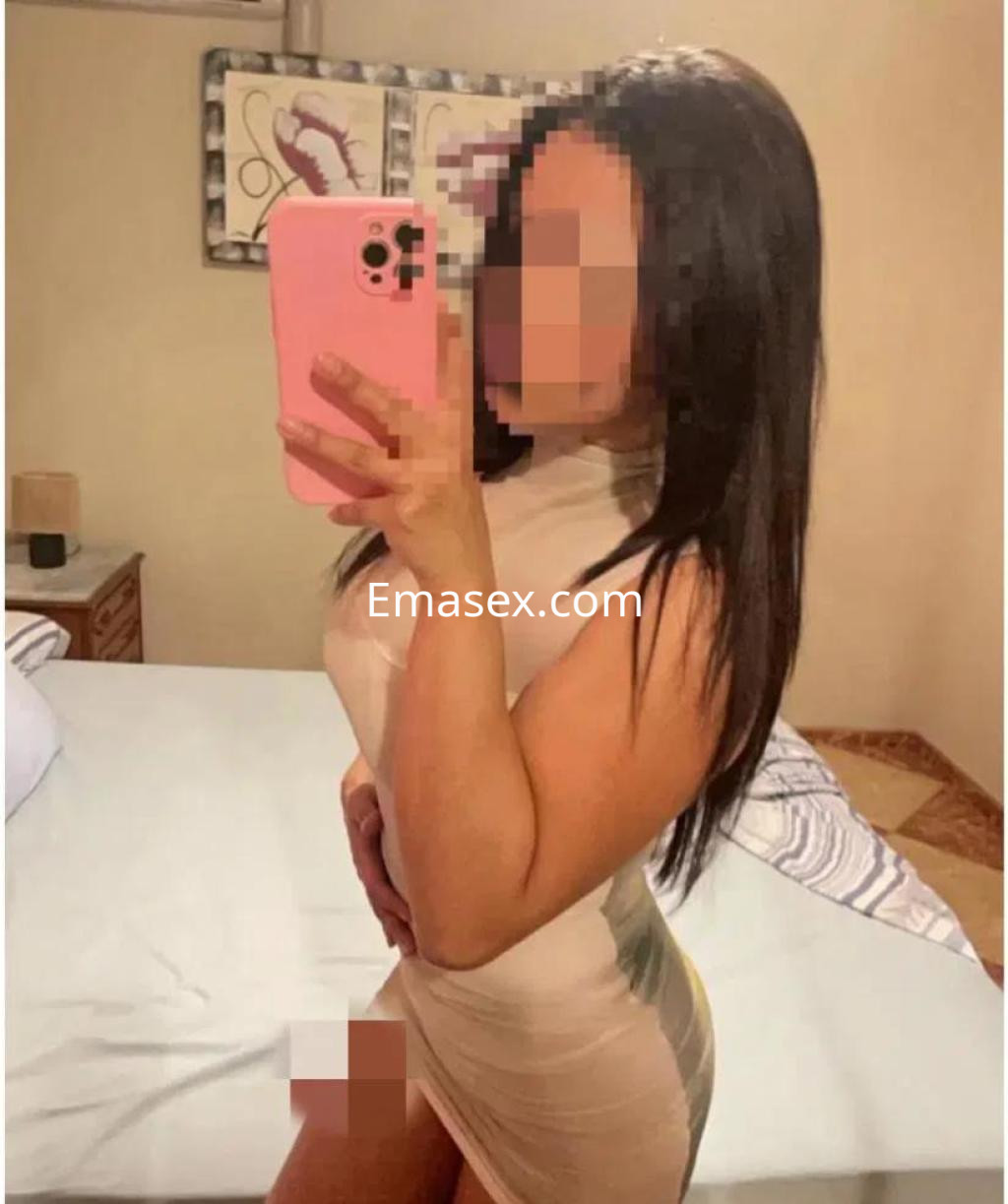 Escort en Albacete posando frente al espejo con un vestido ajustado, cabello largo y lacio, en un entorno de habitación.