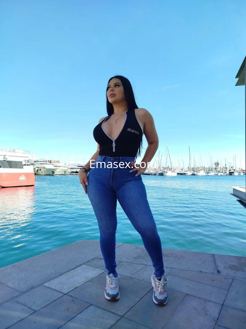 La puta viste un body negro y jeans ajustados, posando con el puerto de Alicante de fondo.