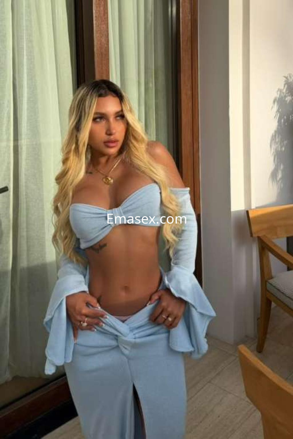 Escort en Ibiza con cabello rubio largo, vistiendo un conjunto de dos piezas azul claro, posando con una expresión seductora.