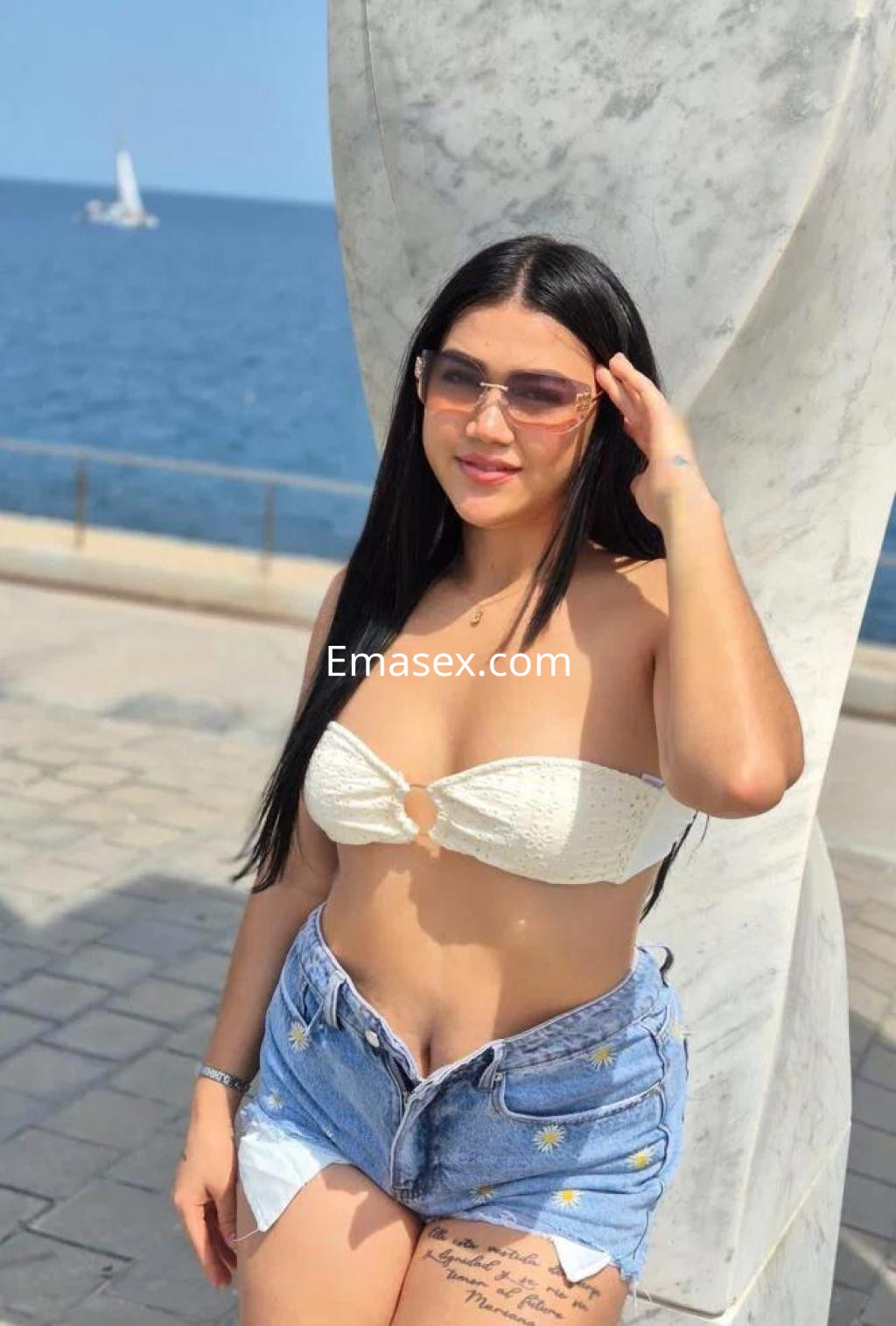Puta posando en Barcelona, lleva un top blanco y shorts de mezclilla, con el mar de fondo y gafas de sol.