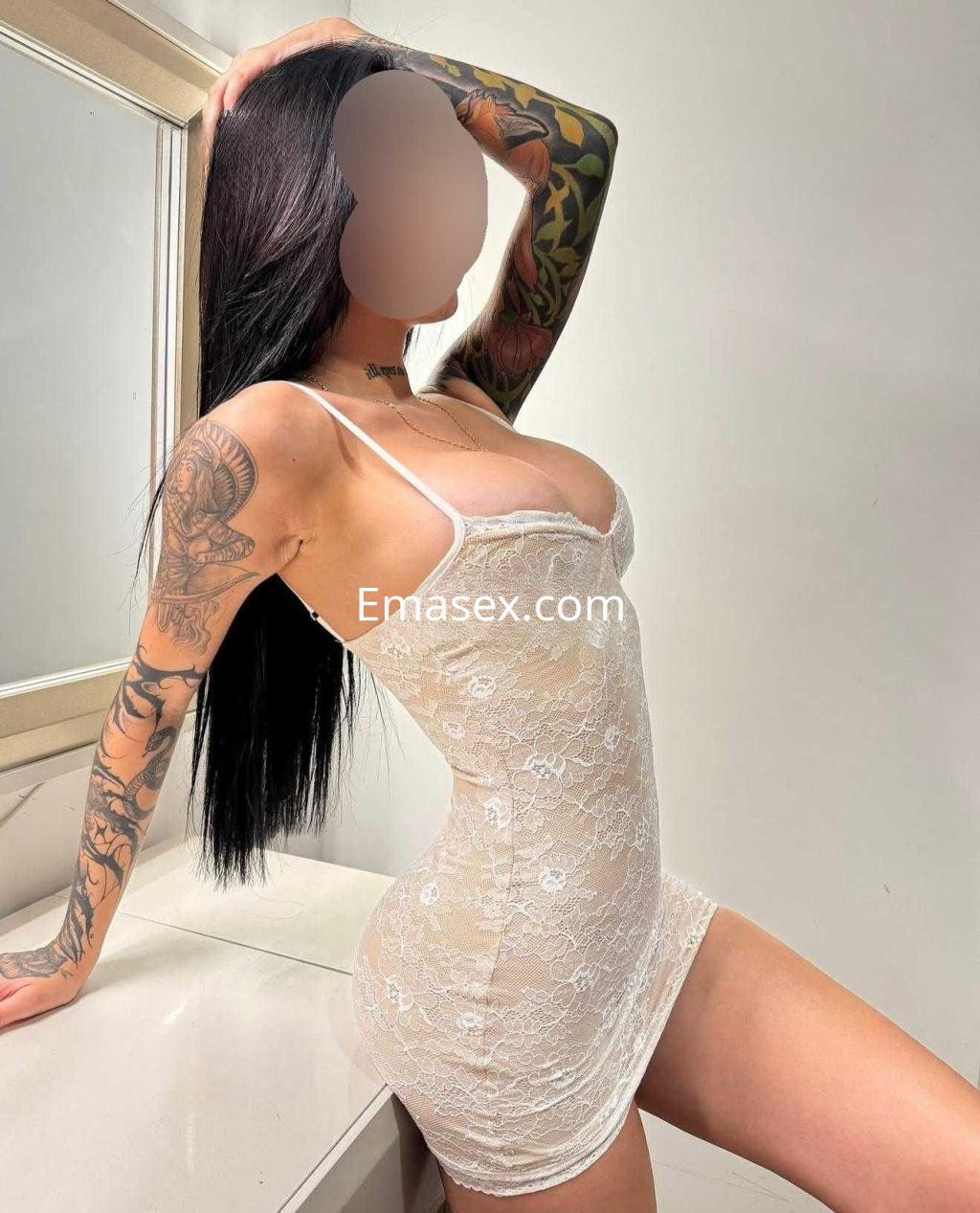 Escort en Barcelona posando de lado con un vestido blanco de encaje, cabello largo y lacio, y tatuajes visibles.