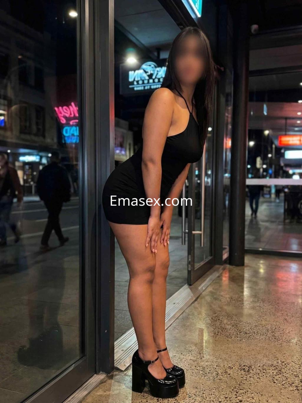 Escort en Barcelona, luciendo un vestido negro ajustado y tacones altos, posando en un entorno urbano nocturno.