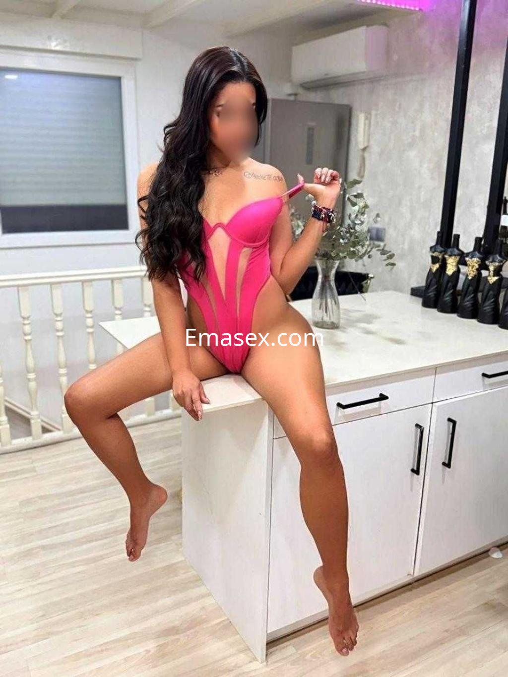Escort en Cádiz posando sobre una encimera, luciendo un bañador rosa con detalles transparentes y cabello largo ondulado.