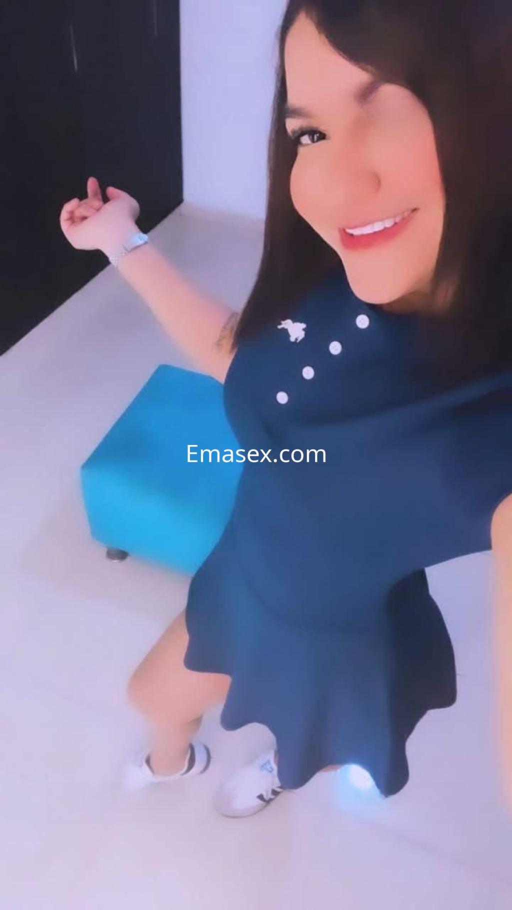 La puta en Málaga luce un vestido azul con detalles blancos y zapatillas deportivas, sonriente en un entorno claro.