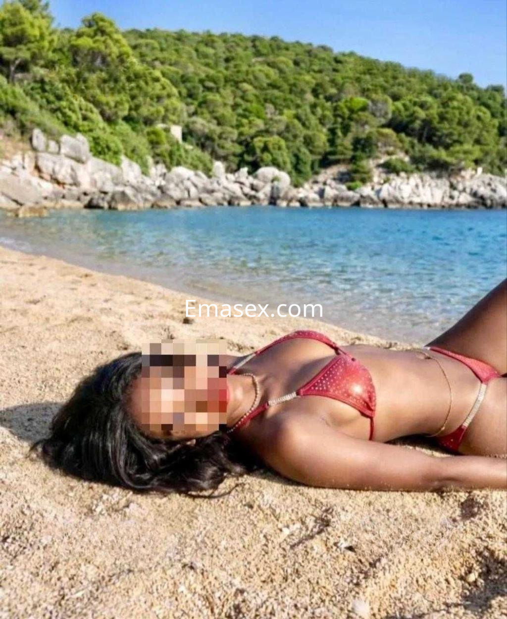 Escort en Zaragoza luciendo un bikini rojo, recostada sobre la arena de una playa con mar azul de fondo.