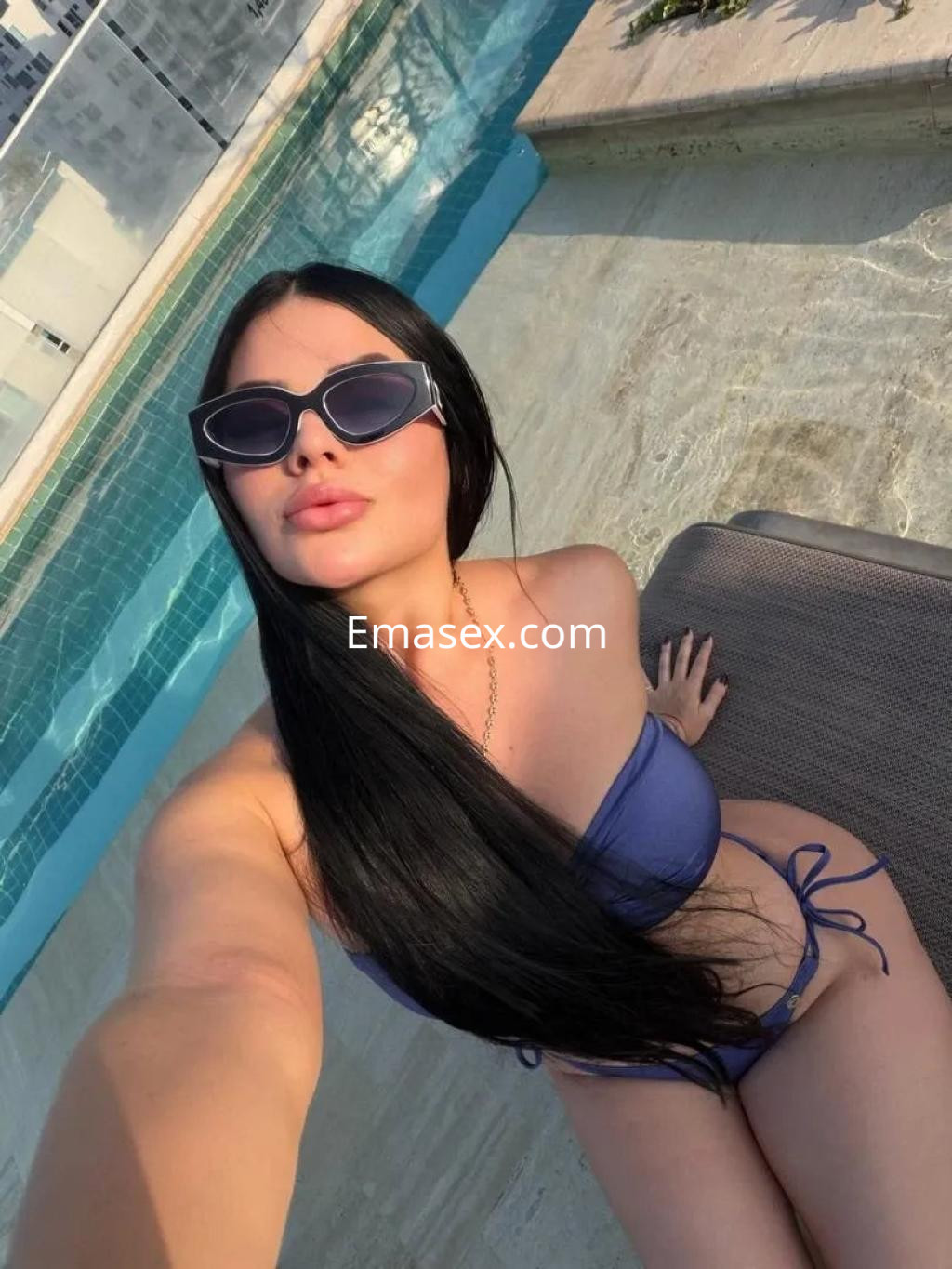 Escort en Zaragoza, con cabello largo y lacio, luciendo un bikini azul y gafas de sol junto a una piscina.