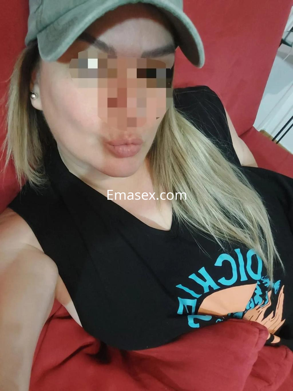 Escort en Zaragoza con gorra, camiseta sin mangas y labios pintados, posando en un sofá rojo.
