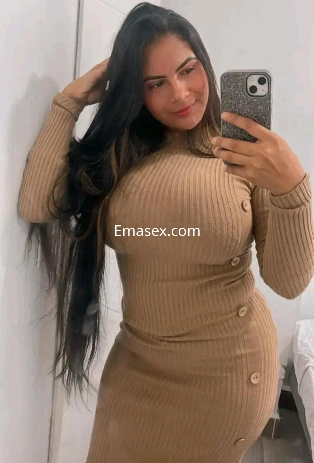 Escort en Granada, con cabello largo y oscuro, viste un vestido ajustado de color marrón claro y posa frente al espejo.