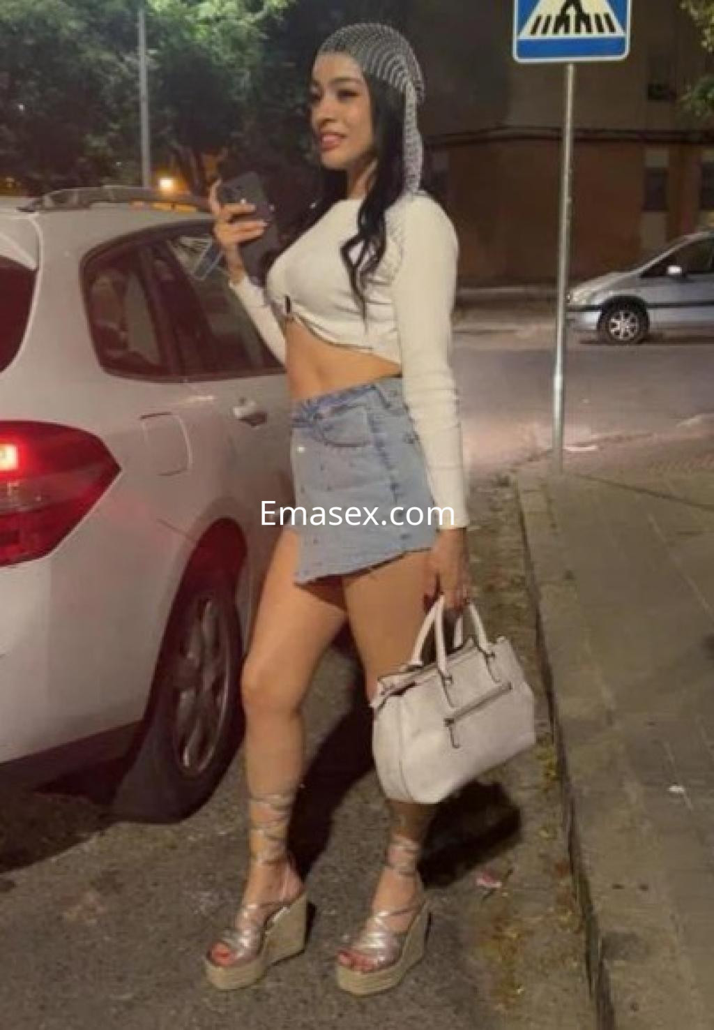 Puta en Sevilla viste un top blanco y una falda de mezclilla, sosteniendo un bolso y un teléfono.