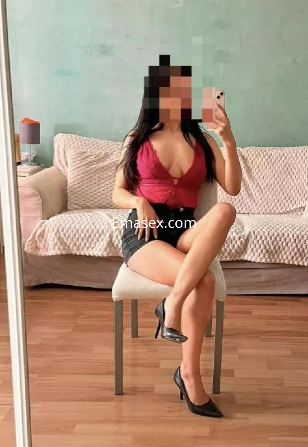 Puta en Madrid sentada en una silla, vistiendo un top rojo y shorts oscuros, con tacones y fondo neutro.
