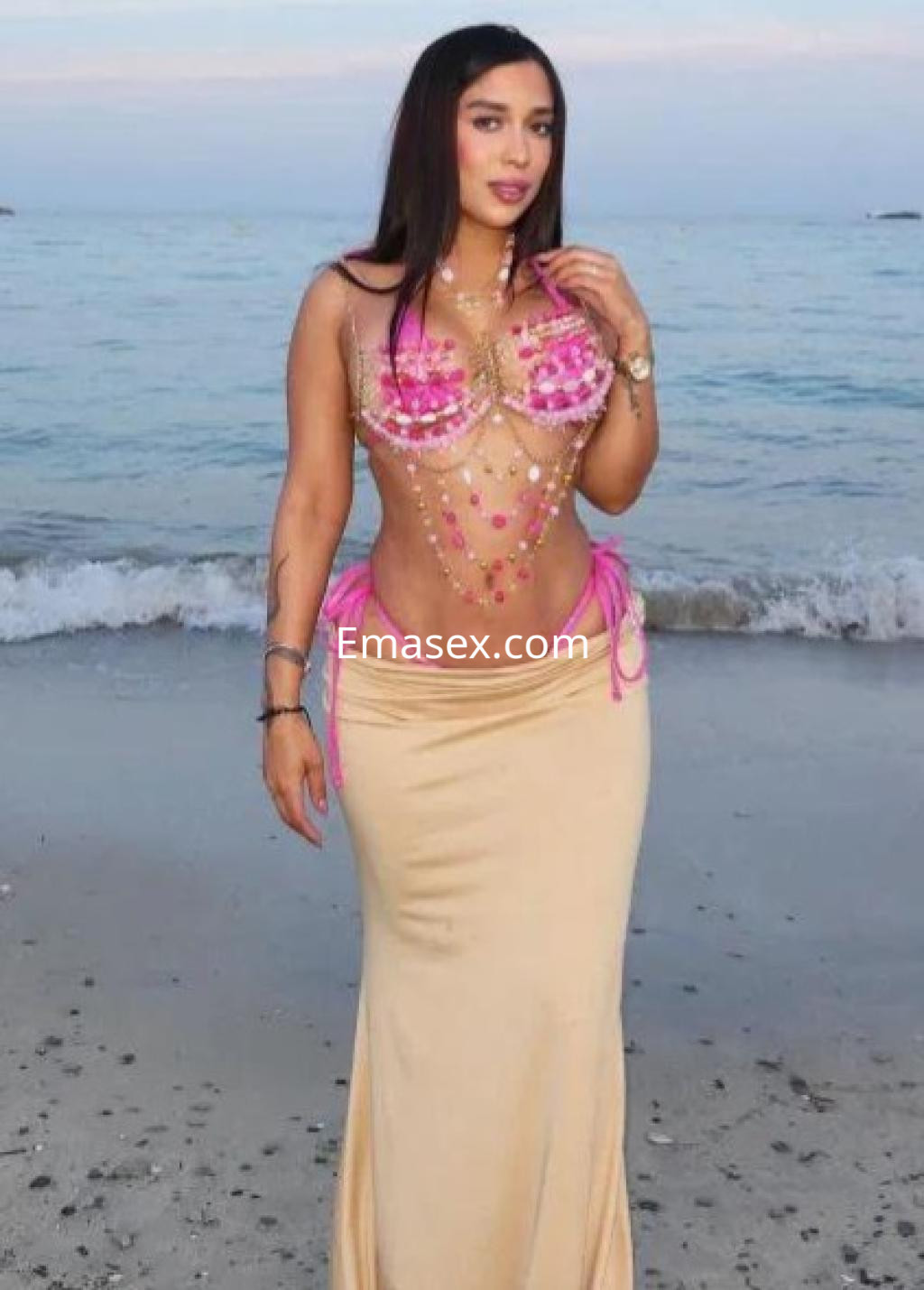 Escort en Girona luciendo un top decorado con perlas y una falda larga, posando junto al mar.