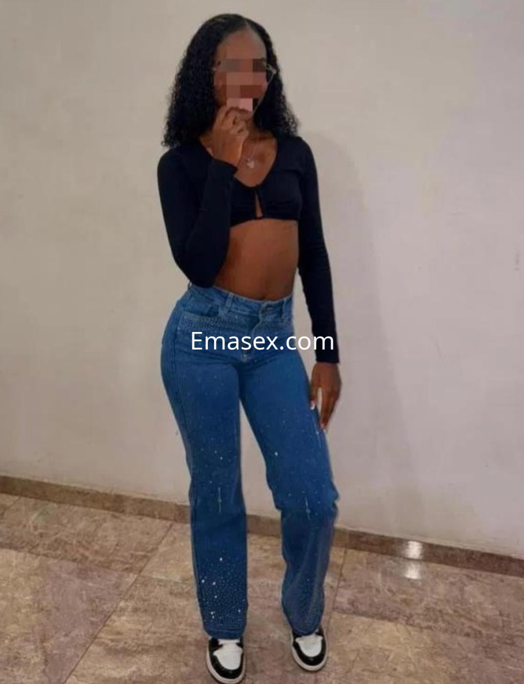 Escort en Castellón, con cabello rizado y oscuro, viste un top negro y jeans claros, posando con confianza.