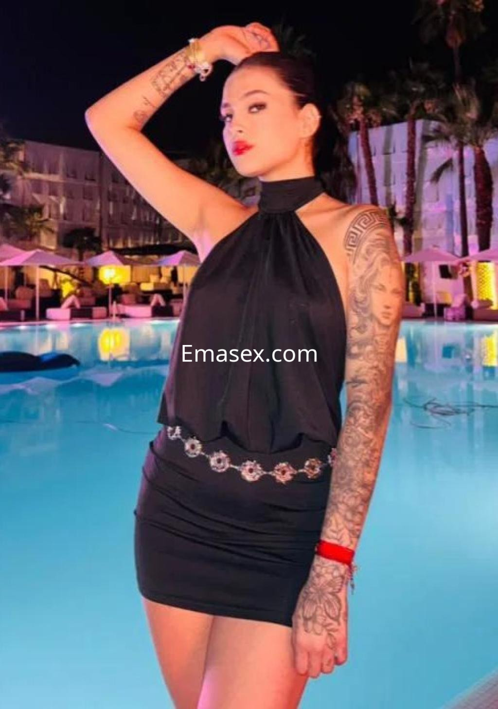 La puta posa junto a una piscina iluminada, luciendo un vestido negro sin mangas y con tatuajes visibles.