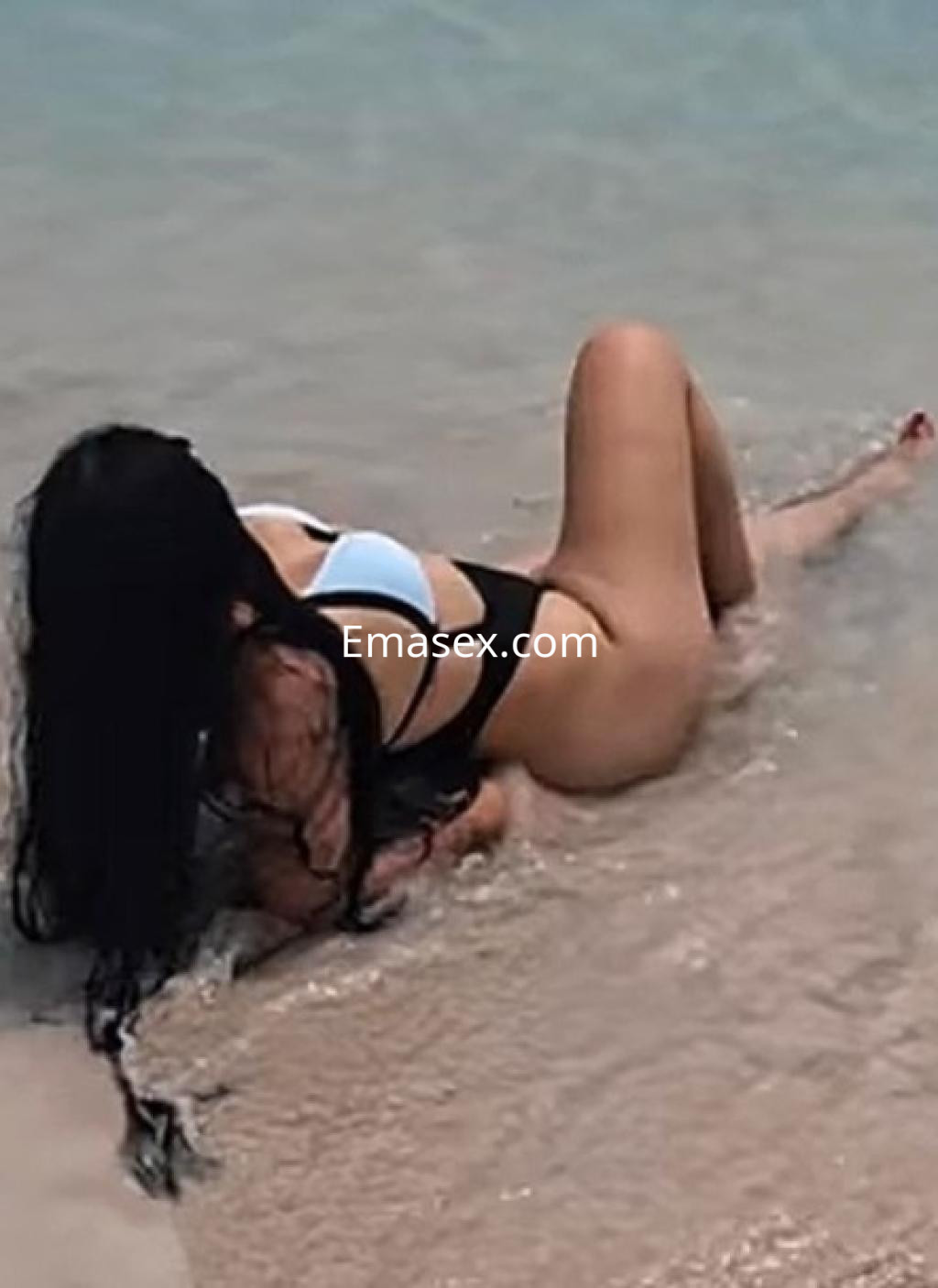 Puta en Huelva, con cabello largo y oscuro, luciendo un bañador negro mientras se relaja en la playa.