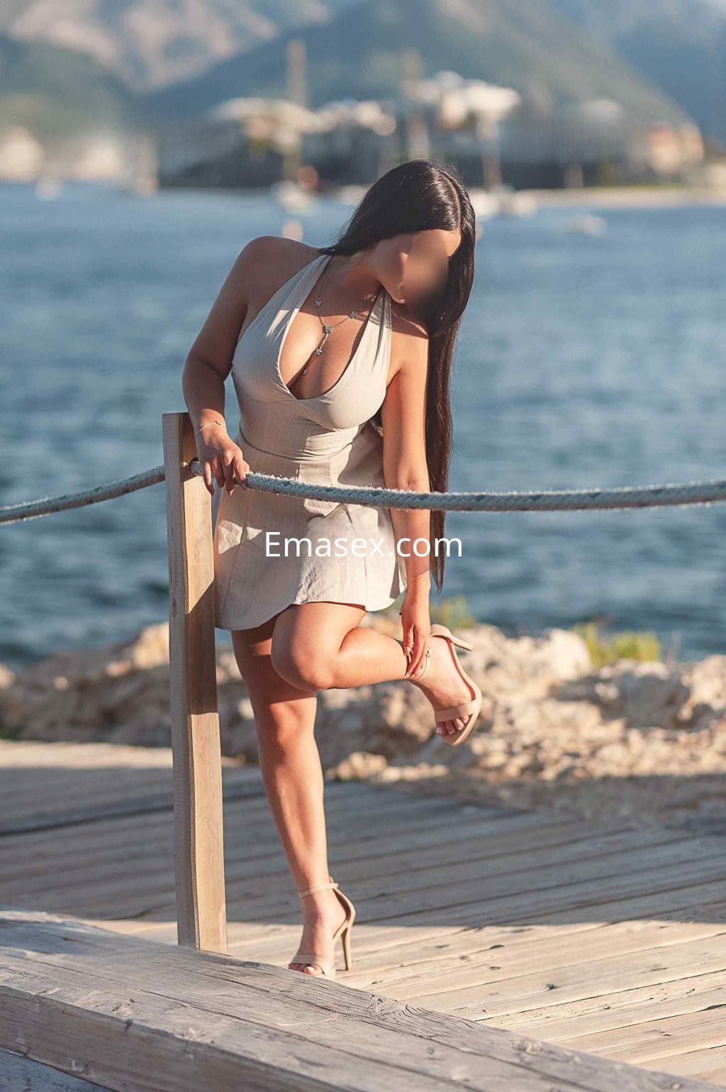 Puta con cabello lacio y oscuro, usando un vestido claro y tacones, posando junto al mar en Alicante.