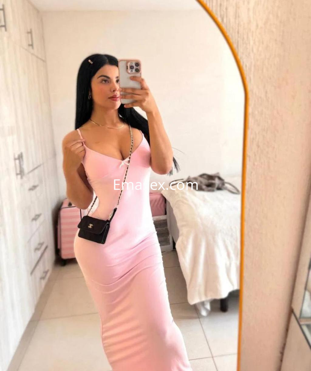 Escort en Alicante con vestido rosa ajustado, posando frente a un espejo en un ambiente interior luminoso.