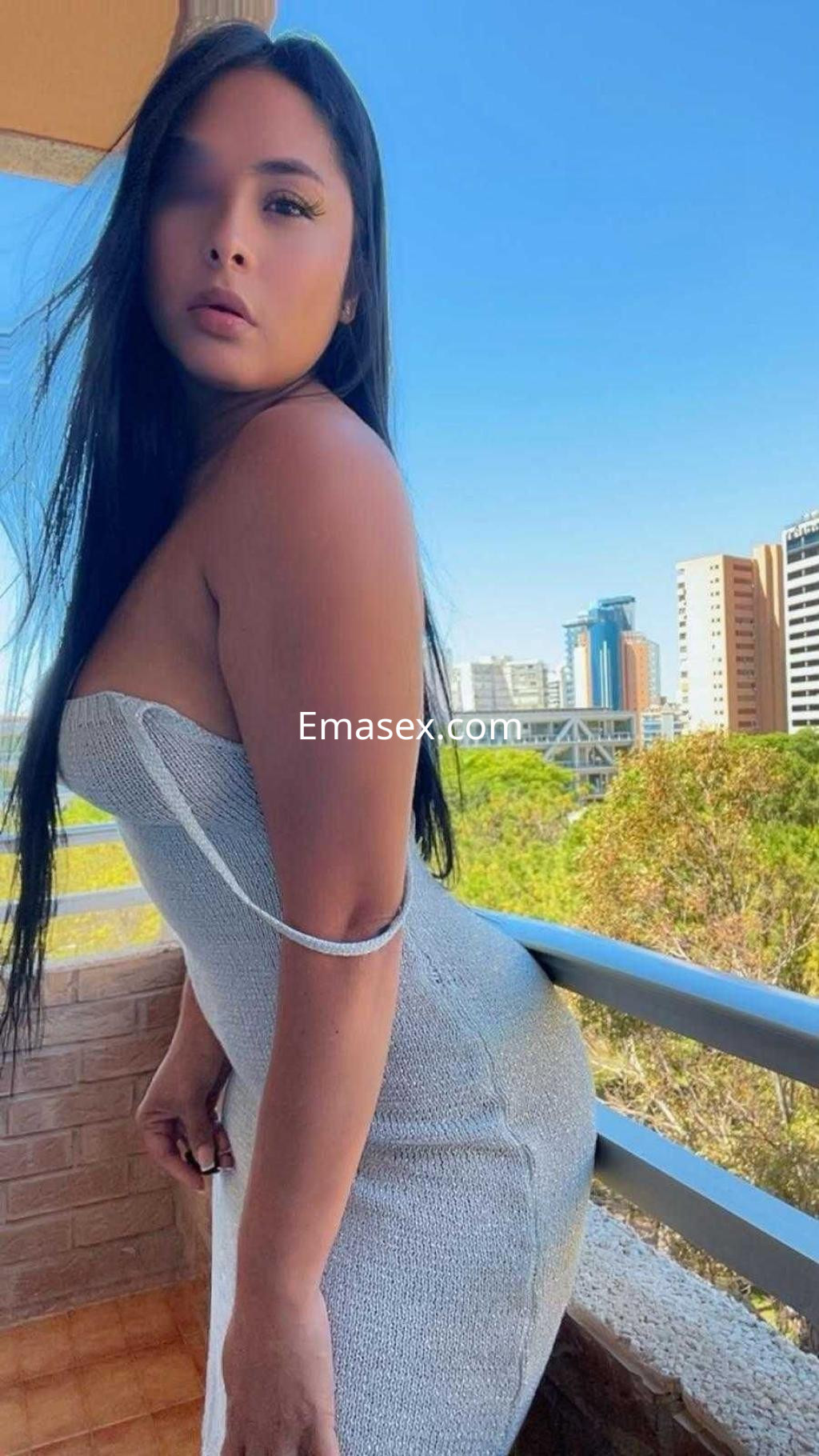 La puta posa con un vestido ajustado en un balcón, mostrando su figura y el paisaje urbano de Alicante.