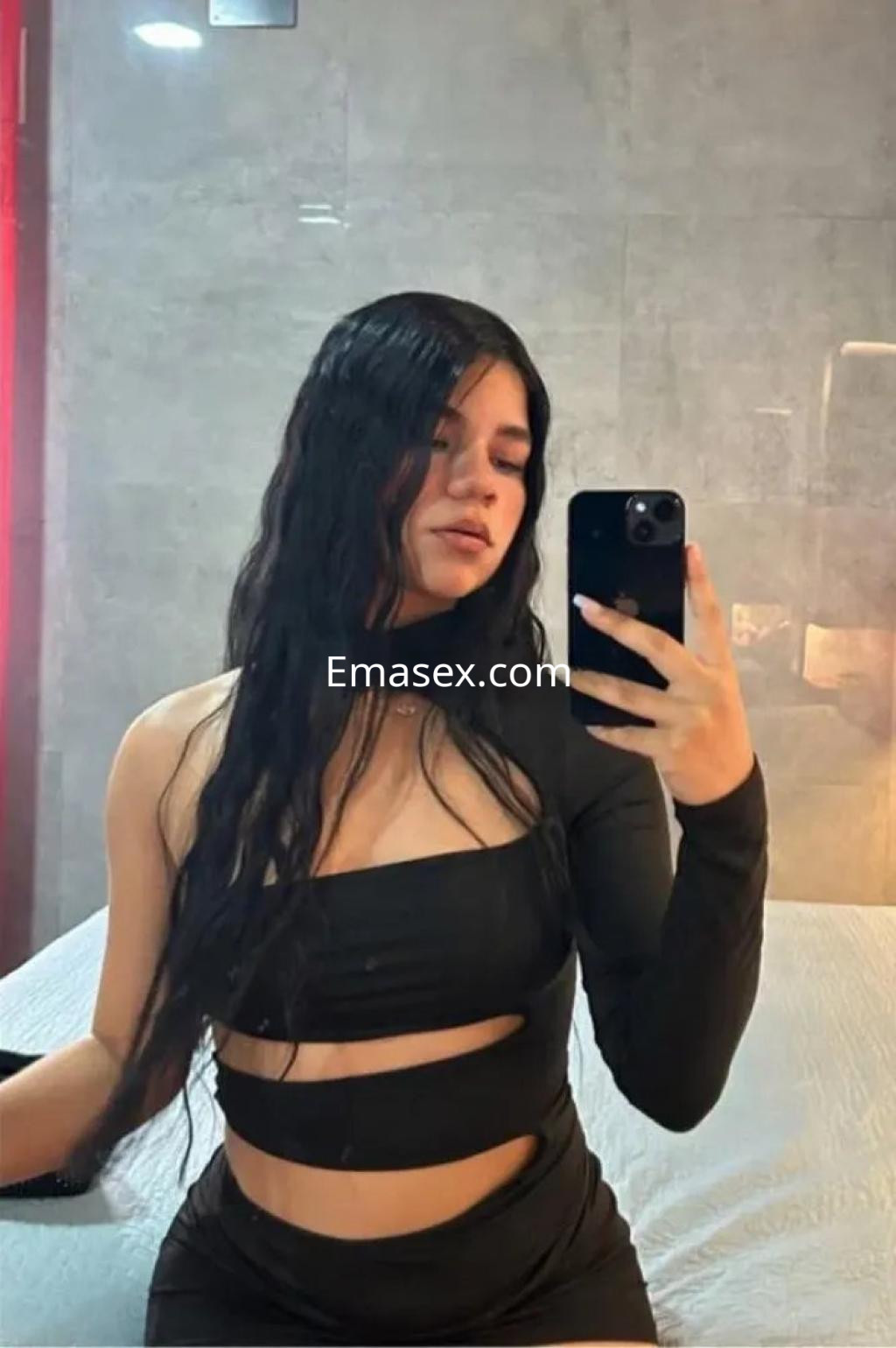 Escort en Alicante con cabello largo y oscuro, vistiendo un top negro asimétrico y posando en un ambiente moderno.