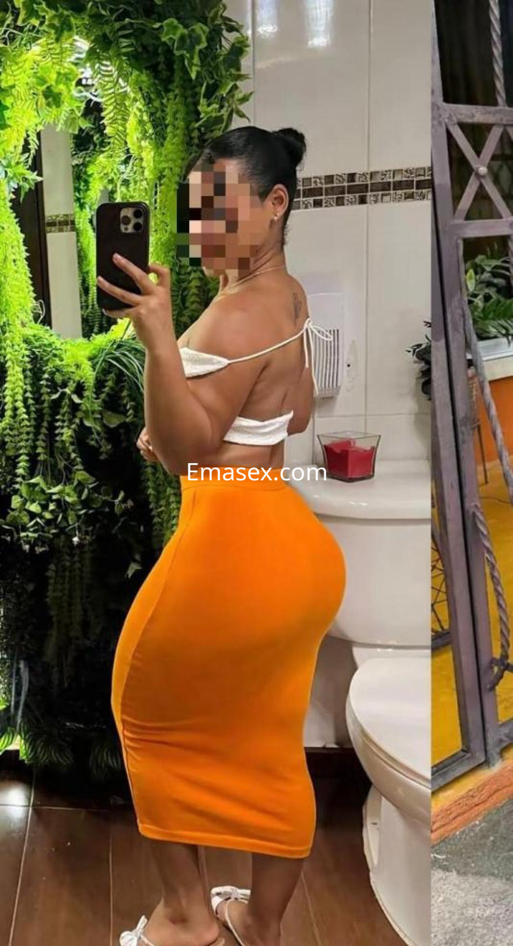 Escort en Almería posando con un vestido naranja ajustado en un baño decorado con plantas verdes.