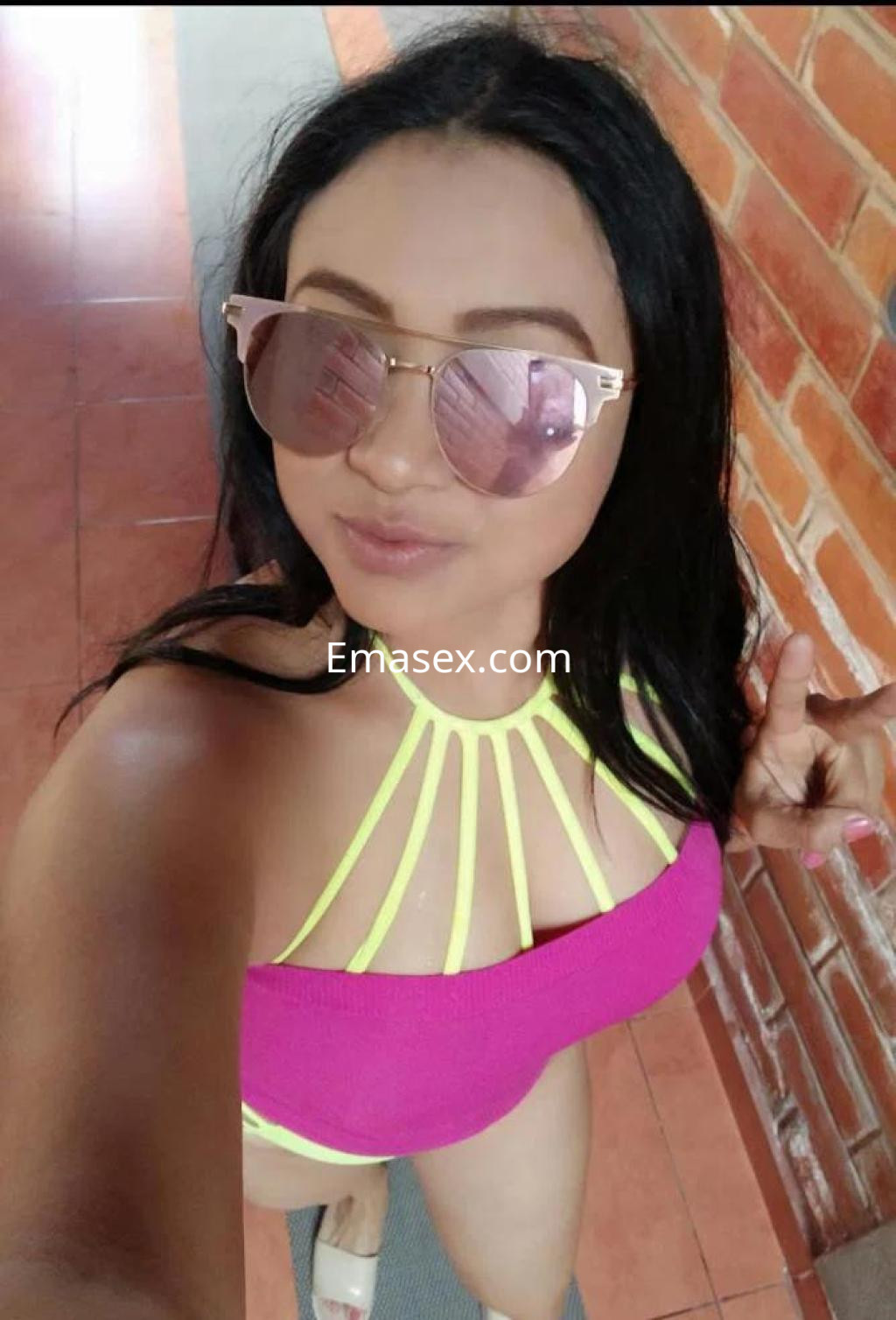 La escort en Almería posa con gafas de sol y un bikini rosa con tiras amarillas, mostrando una sonrisa alegre.