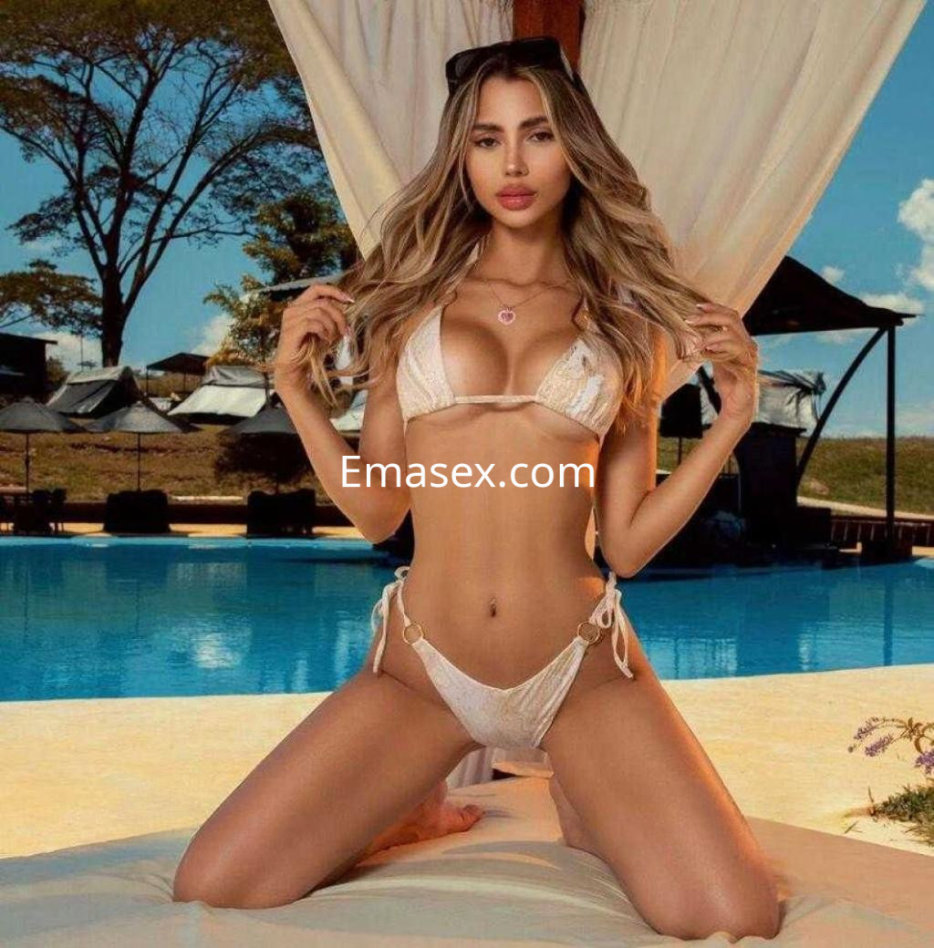 Escort en Barcelona posando en bikini blanco junto a una piscina, con cabello largo y ondulado en un entorno soleado.