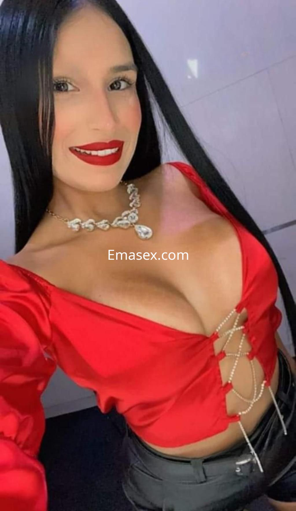 La puta luce un top rojo con escote pronunciado y pantalones oscuros, sonriendo en un ambiente moderno en Cádiz.