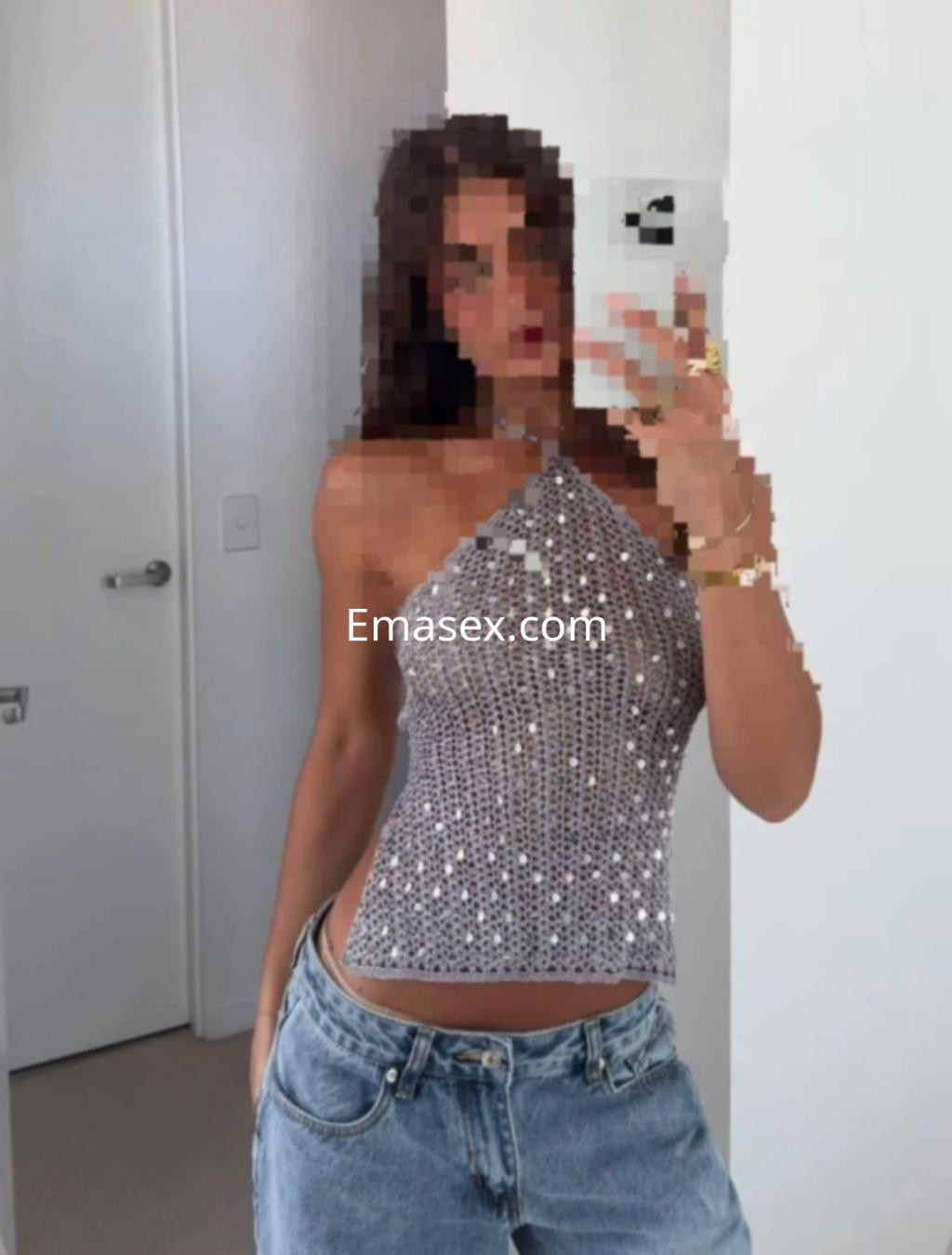 Puta en Cádiz, posando frente al espejo con una blusa brillante y jeans, mostrando una actitud segura.