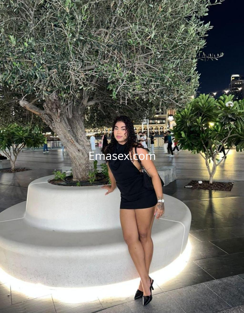 La puta viste un vestido negro ajustado y tacones, posando junto a un árbol en un entorno iluminado en León.
