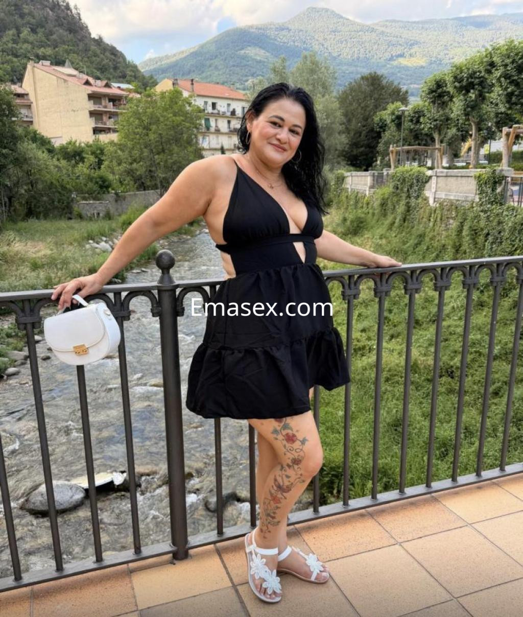 La puta posa con un vestido negro y sandalias blancas en Lleida, frente a un paisaje montañoso.
