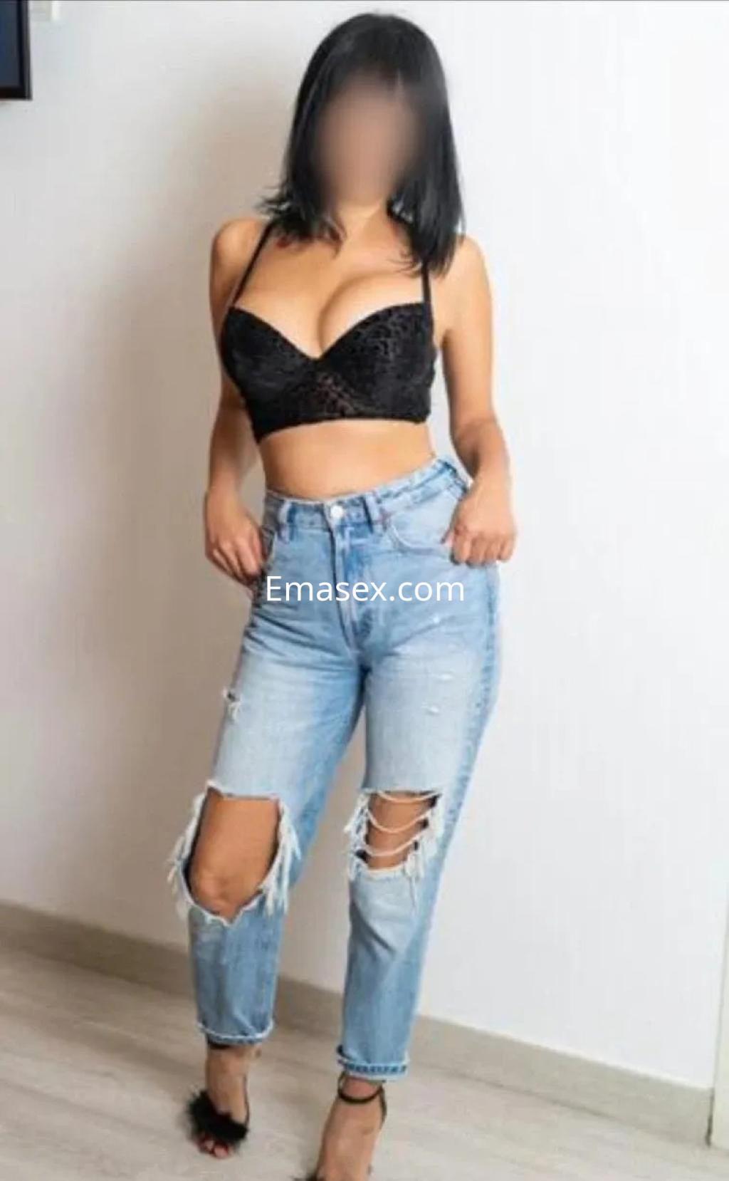 Escort en Lugo con top negro y jeans rotos, posando de pie con una expresión segura en un fondo neutro.