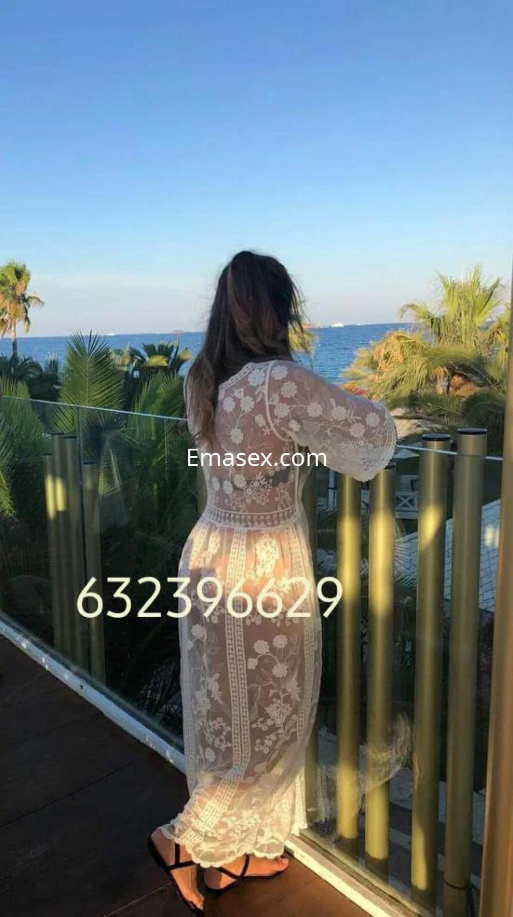 Puta en Murcia, vista desde atrás con un vestido blanco transparente, contemplando el mar y la vegetación.