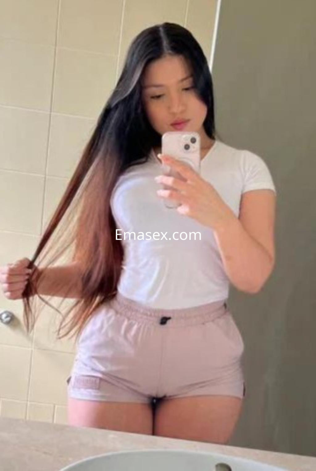 Escort en Sevilla posando en un espejo, viste una camiseta blanca y shorts cortos, con cabello largo y lacio.